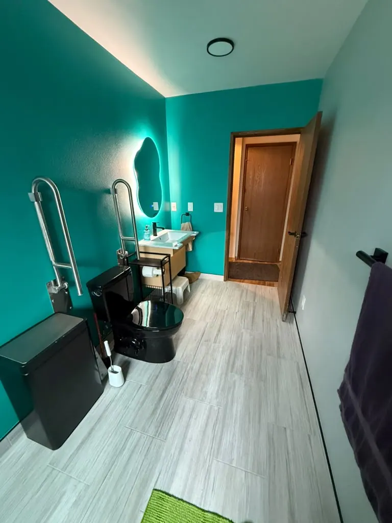 Converted bathroom with a teal accent wall, a black toilet, fold-down grab bars and gray plank tile