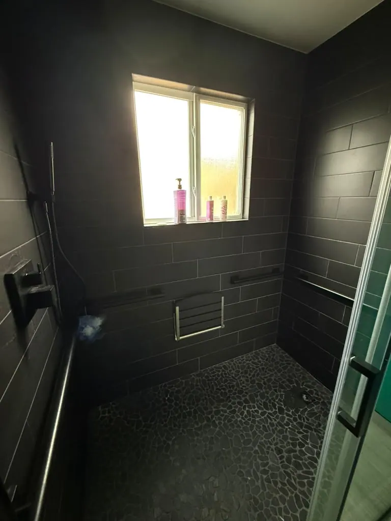 Walk-in shower with dark wall tile, a pebble floor, grab bars and a fold-down shower seat