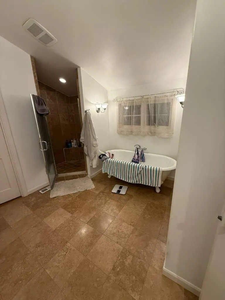 Before: The tub and shower corner before the remodel, with travertine floor tile and a clear glass shower door