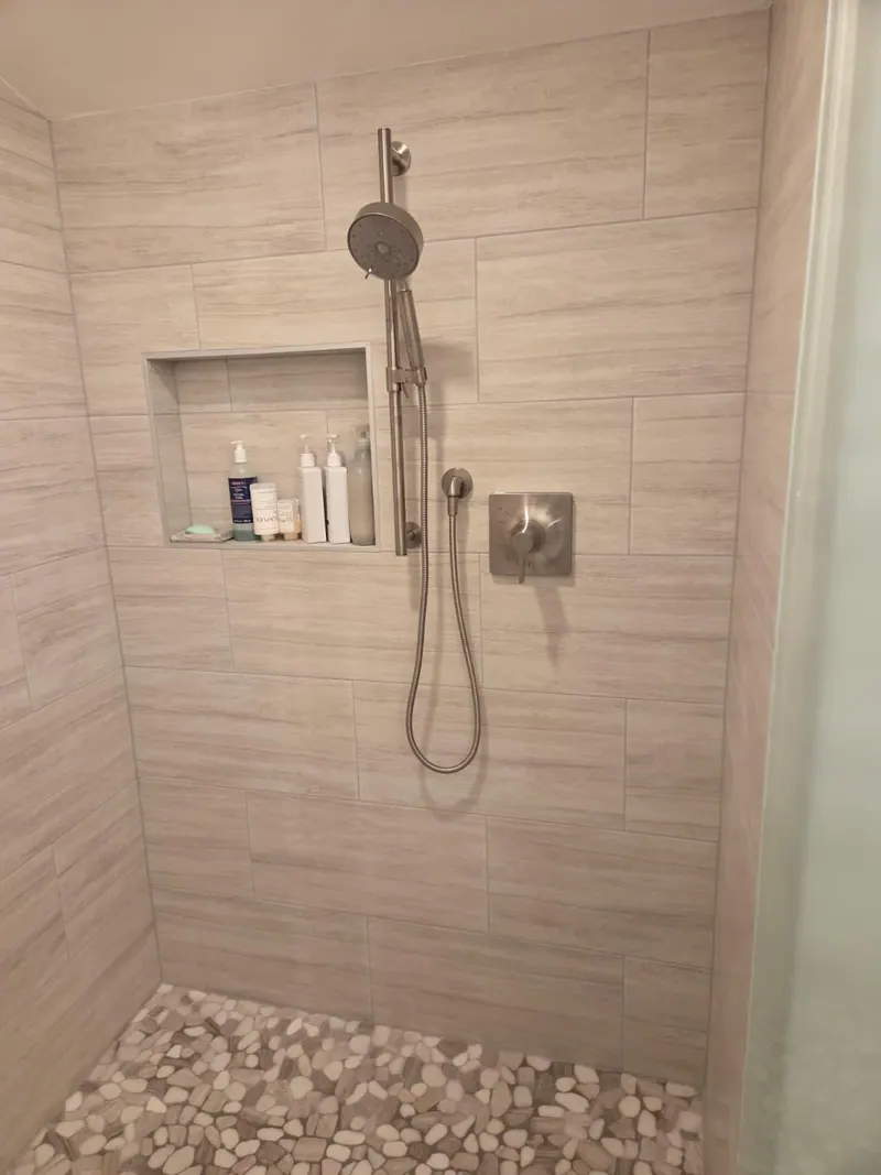 Remodeled walk-in shower with light gray wall tile, a recessed niche, a handheld shower and a pebble floor