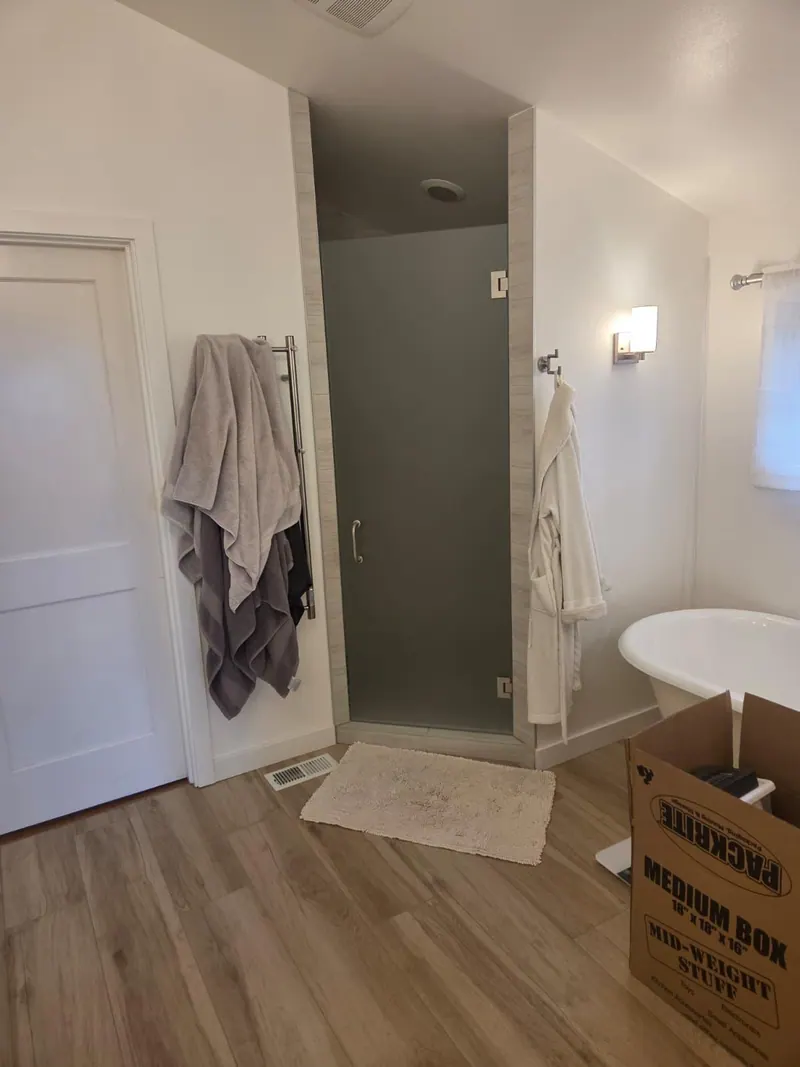 After: Remodeled primary bathroom with a frosted-glass shower door, a freestanding tub and wood-look flooring