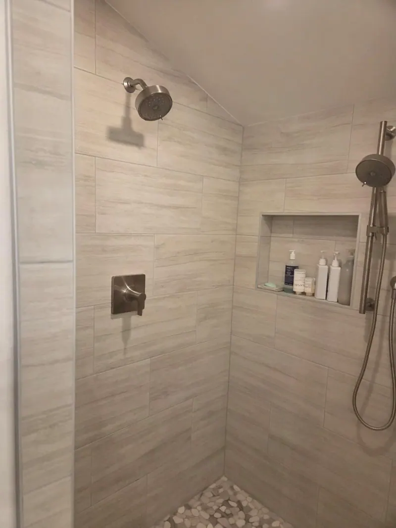 After: Remodeled shower with gray wall tile under a sloped ceiling, a niche, two shower heads and a pebble floor