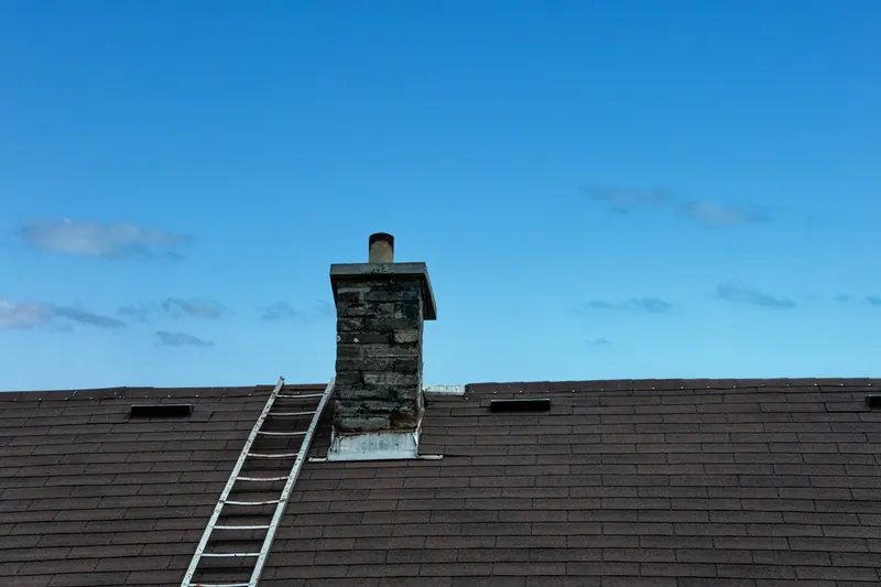 Stone chimney on a shingle roof with a ladder leaning against the roof