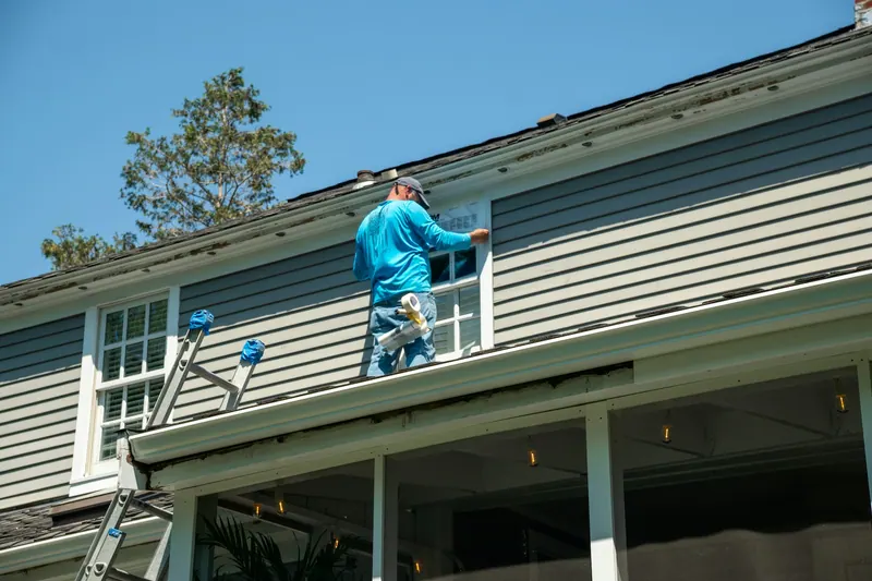 Painter on a ladder working on the trim of a gray-sided house
